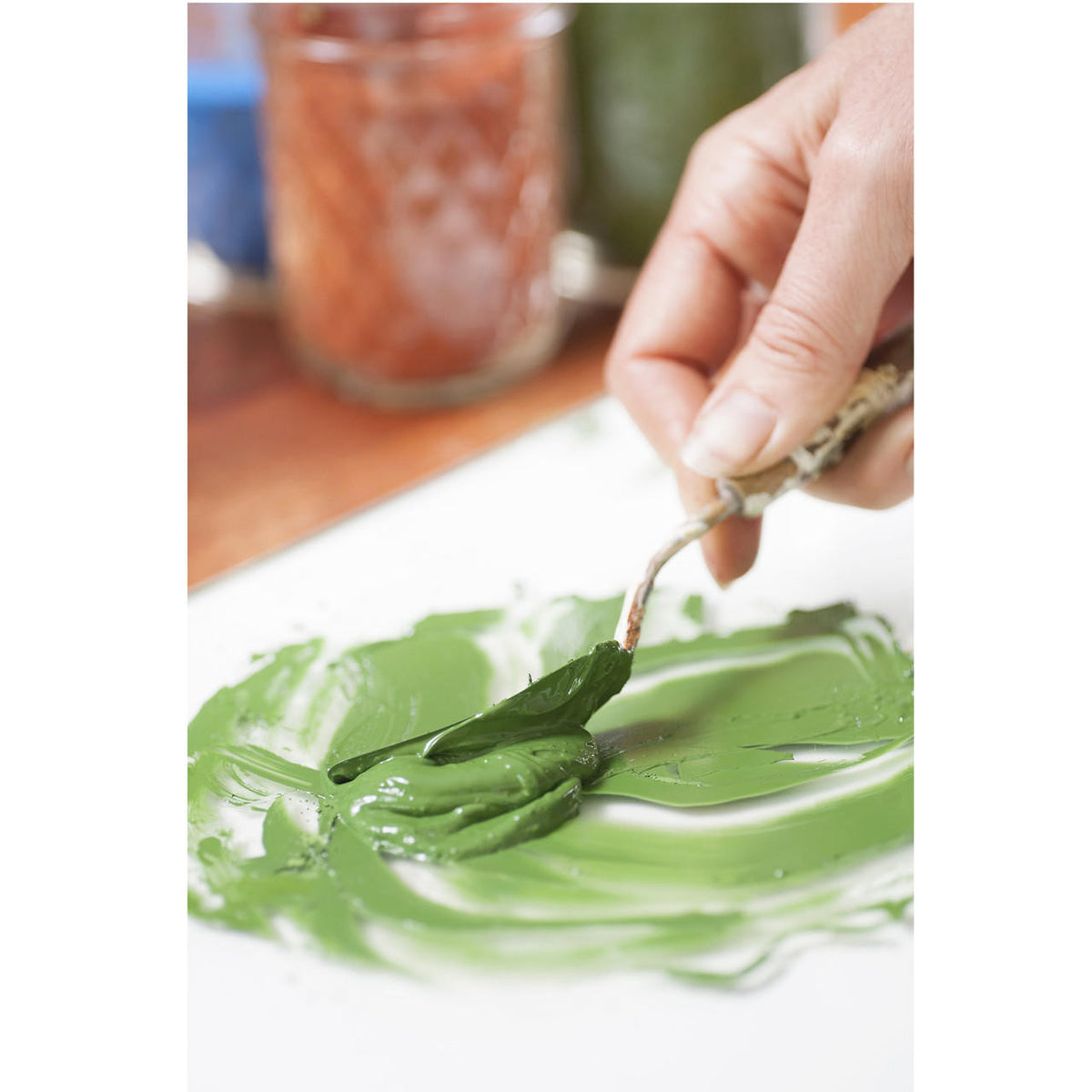 A hand holding a palette knife and mixing green paint on a glass palette