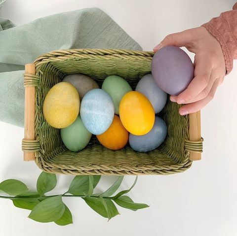 A Basket filled with colorful eggs dyed using Natural Earth Paint's Natural Egg Dye Kit. There is a green cloth fabric in the background along with a green plant branch and a child's hand holding a purple egg.
