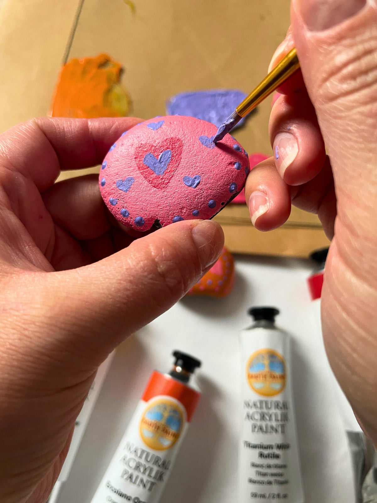 Person holding a rock in one hand using a paint brush with the other hand to paint purple hearts on a pink background using Natural Acrylik Paint