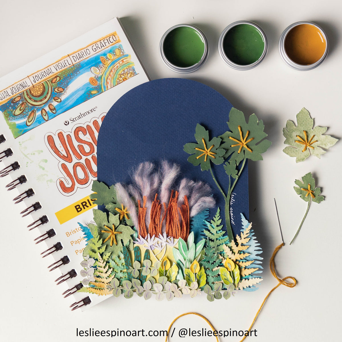 One of Leslie Espino's botanical paper and thread creation in front of their book, "Visual Journal" with 3 containers in the upper right hand corner containing light green, dark green and yelllow ochre paint.  