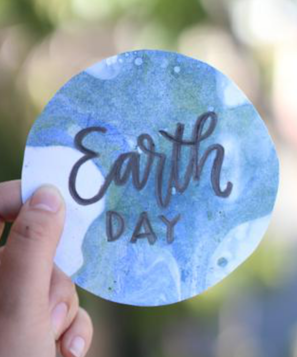 Hand holding up a circle piece of paper that says "Earth Day" in black on a blue and green marbled background that looks like the planet Earth