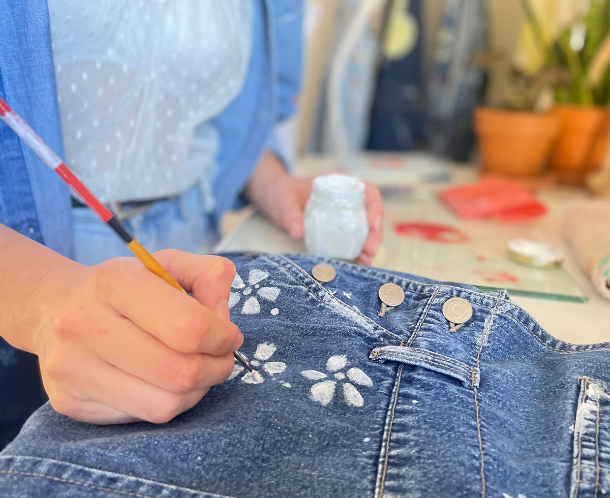 Close up of Artist Micaela Rish's hand painting white flowers on blue jean fabric using a paint brush