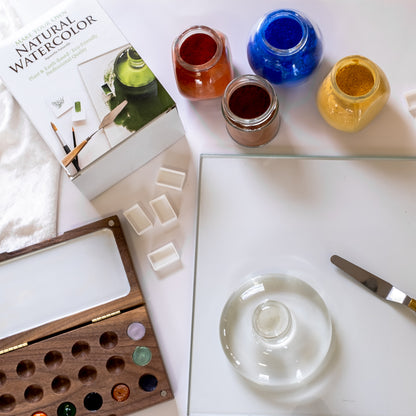 A glass palette, palette knife and glass muller next to four open jars of paint, four empty watercolor pans, a wooden watercolor palette and a Natural Watercolor Kit