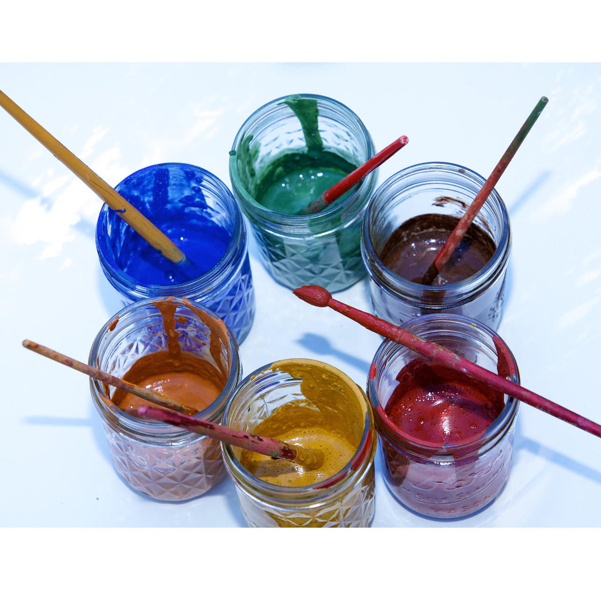 Six open jars of paint with brushes, arranged in a circle