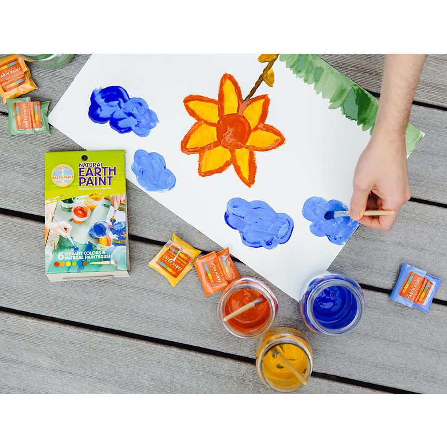 A hand painting a picture on a wooden table top with the contents of the Petite Natural Earth Paint Kit strewn about